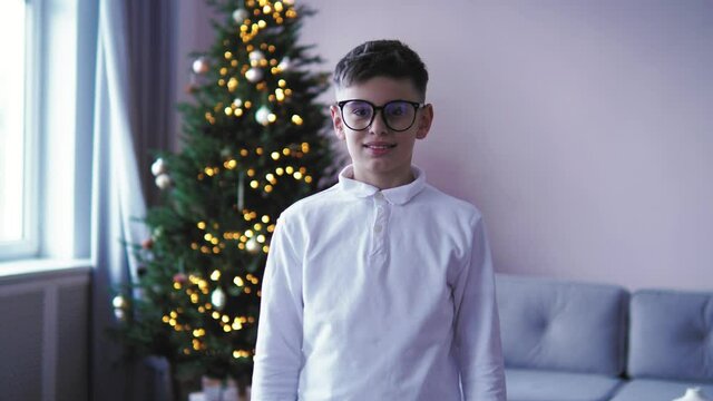 Handsome caucasian smart teenage boy in glasses looking at camera, standing in front of camera, christmas tree with lights on background. Christmas time. Portrait of hispanic kid child boy male 