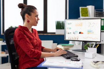 Startup manager in red shirt looking at charts on computer monitor. Smiling small business owner typing on keyboard at desk in start up office. Employee with clipboard analyzing graph and data.