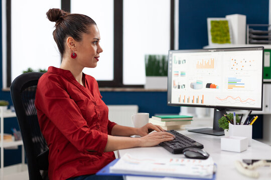 Startup Manager In Red Shirt Looking At Charts On Computer Monitor. Smiling Small Business Owner Typing On Keyboard At Desk In Start Up Office. Employee With Clipboard Analyzing Graph And Data.