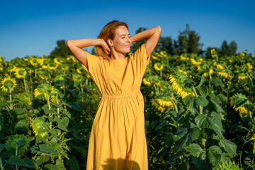 The concept of freedom in a sunflower field. A beautiful relaxed red-haired woman in a yellow dress poses smiling, closing her eyes. Natural feminine beauty © BULAT YAHUDZIN