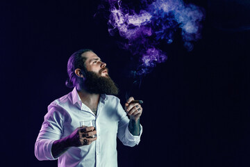 Portrait of handsome bearded man with cigar and glass whiskey while blowing smoke on dark background