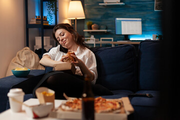Office worker talking on smartphone with friends while holding tasty delicious burger relaxing on couch in living room after work. Woman enjoying phone conversation and having fast food delivery