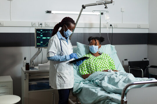 African american therapist doctor with protection face mask against covid19 holding tablet computer with medical expertise explaining healthcare treatment to sick patient. Medicine service