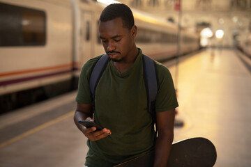 Young man waiting for the train standing at the railway station. Happy guy using the phone while waiting for the train