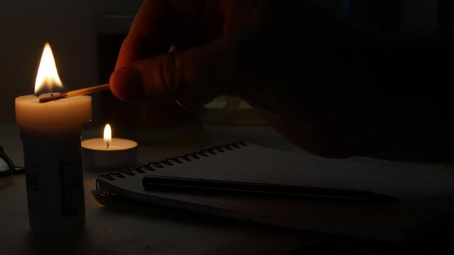 Man Lighting a Candle in a Dark Room and Write with a Pen on a Piece of Paper in Time of an Accidental Power Failure.