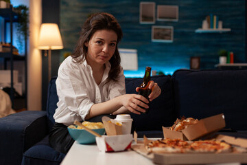 Portrait of smiling woman looking into camera sitting on sofa holding beer bottle in front of coffee table with takeaway dinner. Office worker enjoying a lager and fast food in the evening.