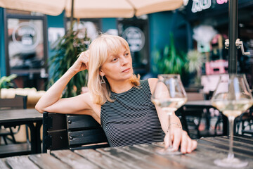 Thoughtful adult mature woman sitting in bar outdoors with wine glasses and blurry restaurant background scene, drinking white wine. Summer sunny day on patio. People lifestyle