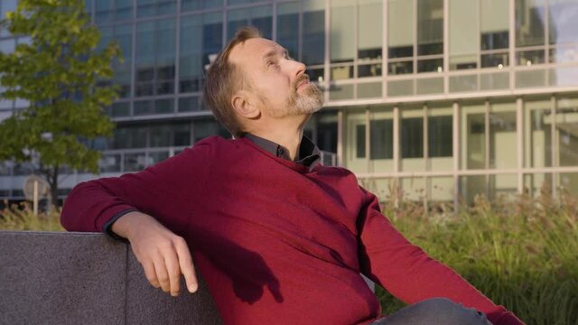 A middle-aged handsome Caucasian man thinks about something as he sits in an urban area - office buildings in the blurry background - closeup