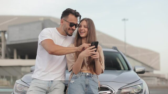 A man and a girl are standing near a car. They are smiling and watching something on their smartphone. The camera is moving around them. 4K