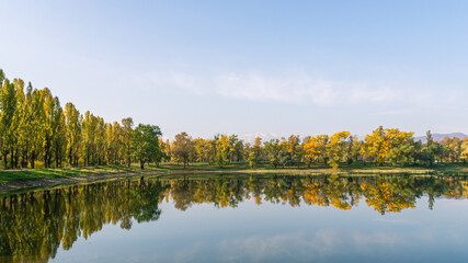 Autumn landscape with a view of the lake, trees and mountains. Reflection of yellowed foliage of trees in the water. Blue sky and mountains in the distance.