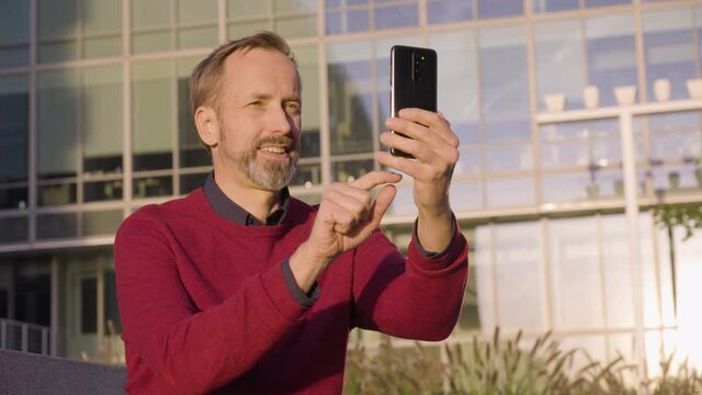 A middle-aged handsome Caucasian man works on a smartphone with a smile as he sits in an urban area - an office building in the blurry background