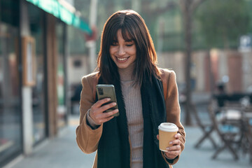 Pretty young woman using her mobile phone while drinking cup of coffee walking the streets of the city.