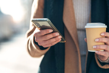 Hands of young woman using her mobile phone while drinking cup of coffee walking the streets of the...