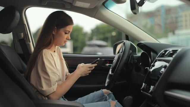 A young woman is sitting in the driver's seat. She is texting on her smartphone. Shooting from the side seats. 4K 50fps
