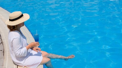 happy senior woman relaxing near blue outdoor swimming pool with blue cocktail wearing straw hat. People are enjoying their summer vacation. All inclusive