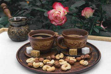 Against the background of roses in the home garden after the rain, on the white surface of the table, there are two clay cups, nuts, white and dark sugar, and a turka. Aromatic coffee crema is visible
