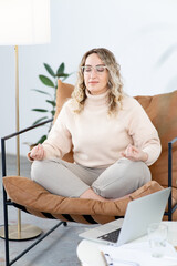 Blonde woman meditates sitting in a chair in the lotus position. Rest from the hard psychological work of a psychotherapist. There is an open laptop on table. There is a large floor lamp nearby.