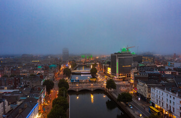 Aerial view of foggy skyline of Dublin with river flowing with bridge connecting two sides of street surrounded by buildings during a cloudy sunrise morning with illuminated lights