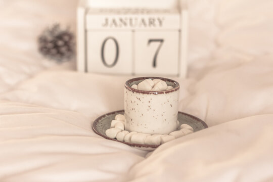 Hot cocoa or coffee with marshmallow in a white ceramic mug, White block calendar present date 7 january on a white blanket. Cozy morning in bed .