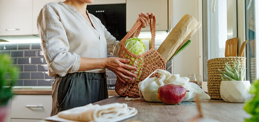 Young woman holding reusable mesh bag with products in the kitchen