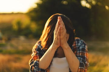 Young woman closed her eyes, praying in a field during beautiful sunset. Hands folded in prayer...