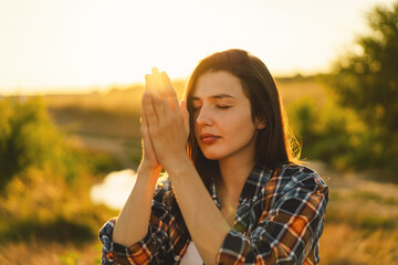 Young woman closed her eyes, praying in a field during beautiful sunset. Hands folded in prayer concept for faith, spirituality and religion. Peace, hope, dreams concept - obrazy, fototapety, plakaty