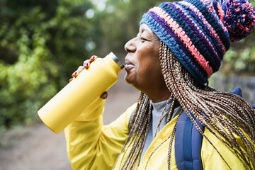 Senior african woman drinking water during trekking day in mountain forest - Focus on hat - Powered by Adobe