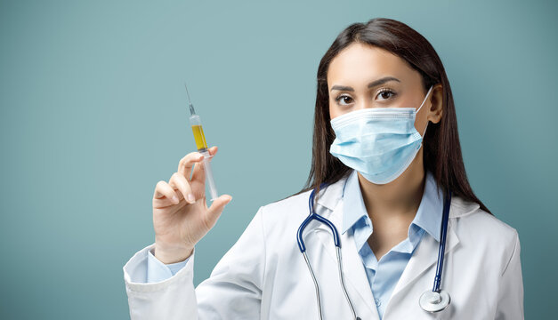 Portrait of female doctor wearing protective mask and gloves holding a syringe isolated on coloured background, studio shot.