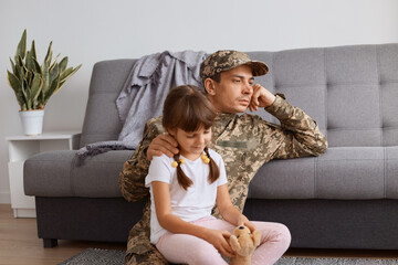 Indoor shot of pensive Caucasian soldier man wearing camouflage uniform returning home after army, spending time with his little daughter and looking away with thoughtful facial expression.