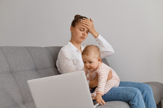 Indoor shot of tired woman with dark hair and bun hairstyle, sitting on sofa with infant baby daughter, looking at laptop, keeping hand on forehead, looks exhausted. - Powered by Adobe