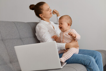 Indoor shot of exhausted female with bun hairstyle wearing white shirt and jeans sitting on sofa and stretching arms after working on laptop, holding crying baby girl.