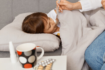 Profile portrait of little sick girl lying on sofa, mother giving her spoon with syrup, kid having...