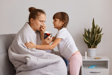 Indoor shot of unhealthy woman drinking tea and suffering runny nose, cute child hugging sick...
