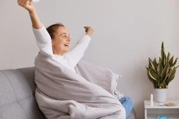 Fotobehang Indoor shot of happy cute girl sitting on sofa and enjoy about getting better after an illness, recovery after disease, cold or flu, healthcare, satisfied female with raised hands. © sementsova321