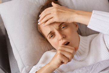 Fototapeta na wymiar Horizontal shot of sick woman with high temperature coughing and touching her head, having influenza symptoms, female lying on sofa, looking away, health care.