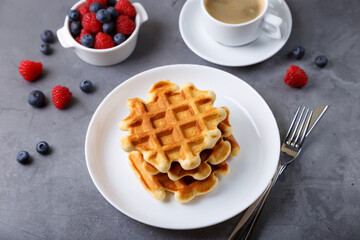 Viennese or Belgian waffles with fresh berries (raspberries and blueberries) on a white plate and a cup of coffee. Traditional dessert. Close-up, grey background, top view. - obrazy, fototapety, plakaty
