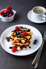 Viennese or Belgian waffles with fresh berries (raspberries and blueberries) on a white plate and a cup of coffee. Traditional dessert. Close-up, grey background.