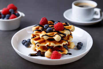 Viennese or Belgian waffles with fresh berries (raspberries and blueberries) on a white plate and a cup of coffee. Traditional dessert. Close-up, grey background.