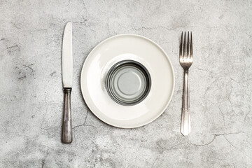 Bote de conserva vacío sobre un plato blanco junto a un tenedor y cuchillo sobre un fondo gris de...