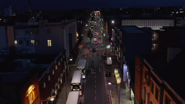 Forwards tracking of group of cars driving in one way street in night city. Road lined by parked vehicles and townhouses. Limerick, Ireland