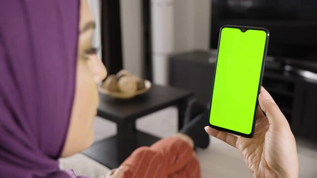 A Muslim woman looks at a smartphone with green screen in an apartment - closeup from behind