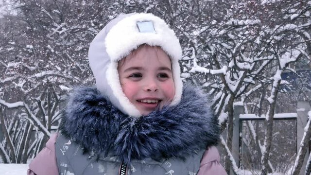 Portrait of little girl in frosty winter outdoors in snowfall. The child is warmly dressed in a gray fur collar, a hat with earflaps with white fur.