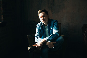 Portrait of Young stylish man in denim or jeans clothes who seat on the armchair in photo studio.