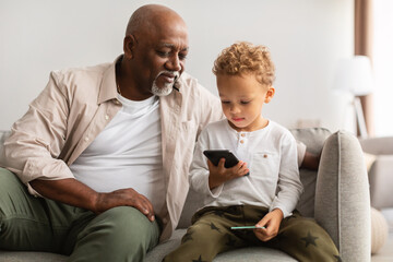 Black Grandfather And Grandson Using Cellphone Sitting On Couch Indoor