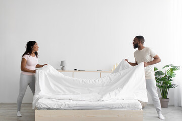 Cheerful young black woman and guy make bed and hold blanket in bedroom interior