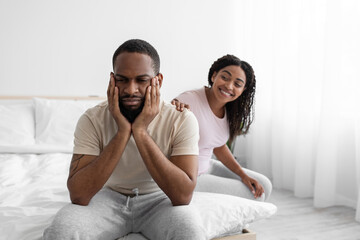 Smiling pretty young black wife calm to sad husband, sit on bed in bedroom interior