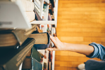 Dark haired woman visitor hand takes large old textbook with hard covering from shelf in spacious modern public library close view from above