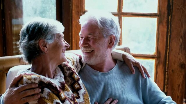 Young senior happy couple sitting together on a bamboo chair at home in wooden chalet enjoying winter or autumn season with love and friendship. Old mature people lifestyle