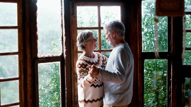 Happy senior couple in love dance at home agains a big windows view with nature woods outside. Romance leisure activity mature man and woman elderly retired lifestyle