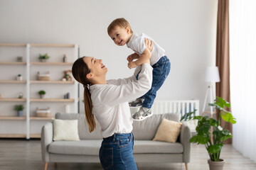 Laughing young mother playing with cute toddler son, holding funny kid pretending flying, having fun together at home